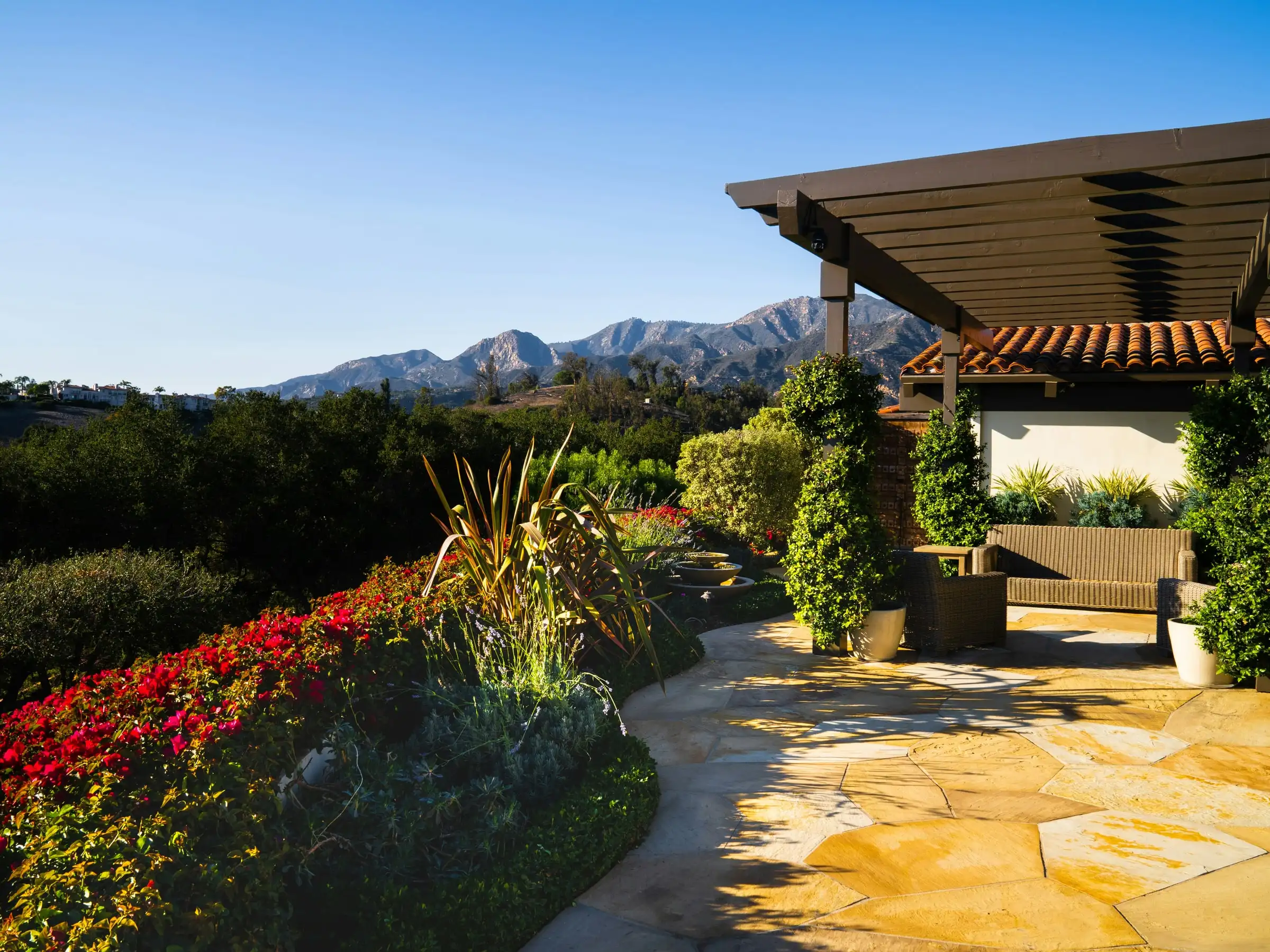A flagstone terrace under a timber pergola, edged with red flowers, mountains behind