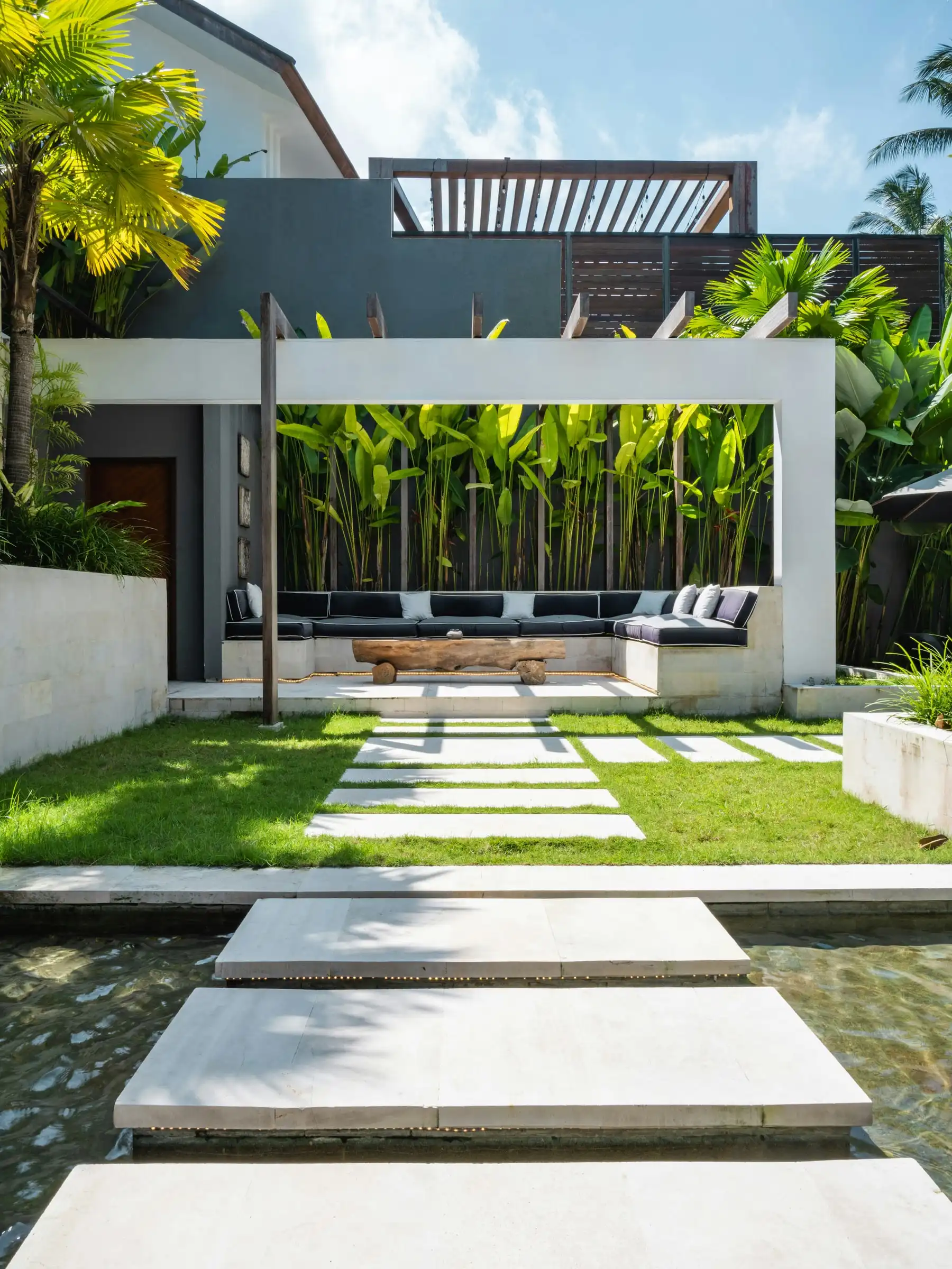 A white pergola frame over a built-in seat, with a wall of tall leaves behind and stepping pavers across the lawn