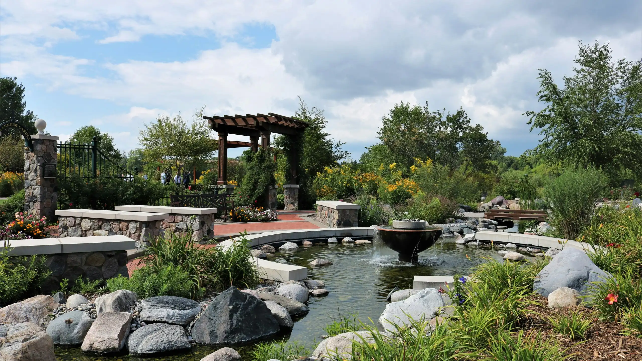 A timber pergola beyond a round pond with a bowl fountain, edged with boulders, stone seats and planting
