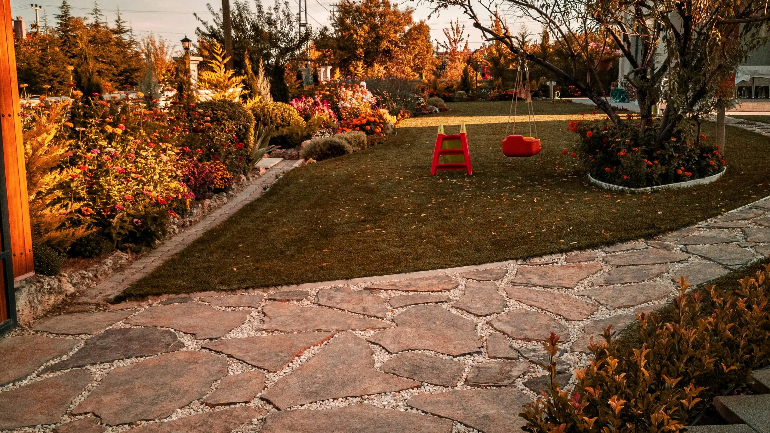 A flagstone path beside a lawn with two red swings hung from a tree, garden beds along the fence
